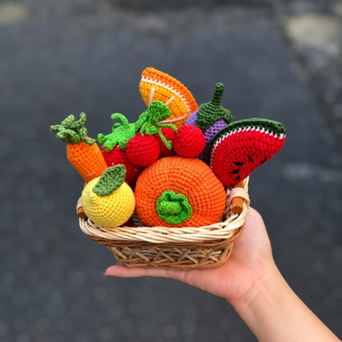 A hand holding a small crochet basket filled with tiny fruit and vegetables