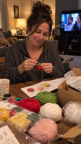 Laura crocheting a strawberry at her table, yarn in an open box and the TV on behind her